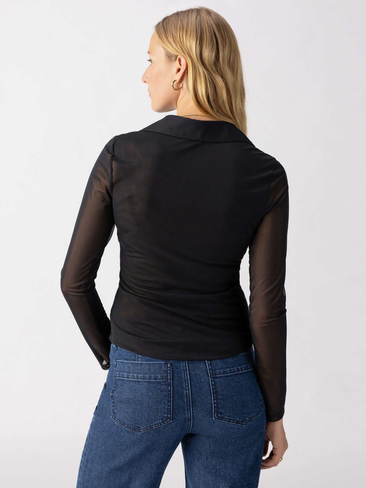 A person with long blonde hair is facing away from the camera, wearing the Dreamgirl Mesh Button Up Black by Sanctuary Clothing and blue jeans. The background is plain and light-colored.