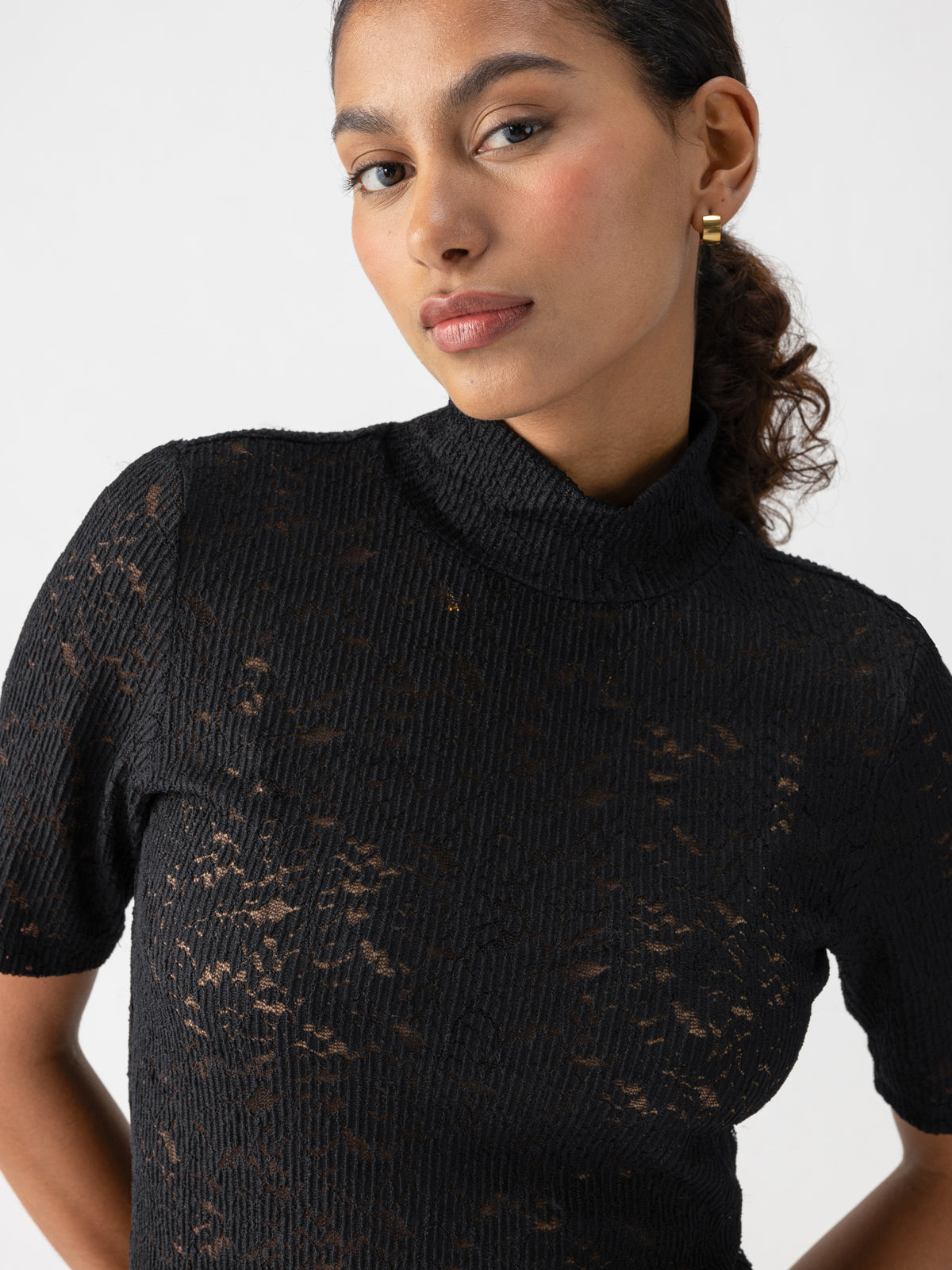 A person with dark, curly hair tied back wears the Sanctuary Clothing S S Lace Mock Black turtleneck top with short sleeves. The semi-see-through textured top reveals a hint of skin and a light undergarment. They are looking slightly off to the side with a neutral expression.