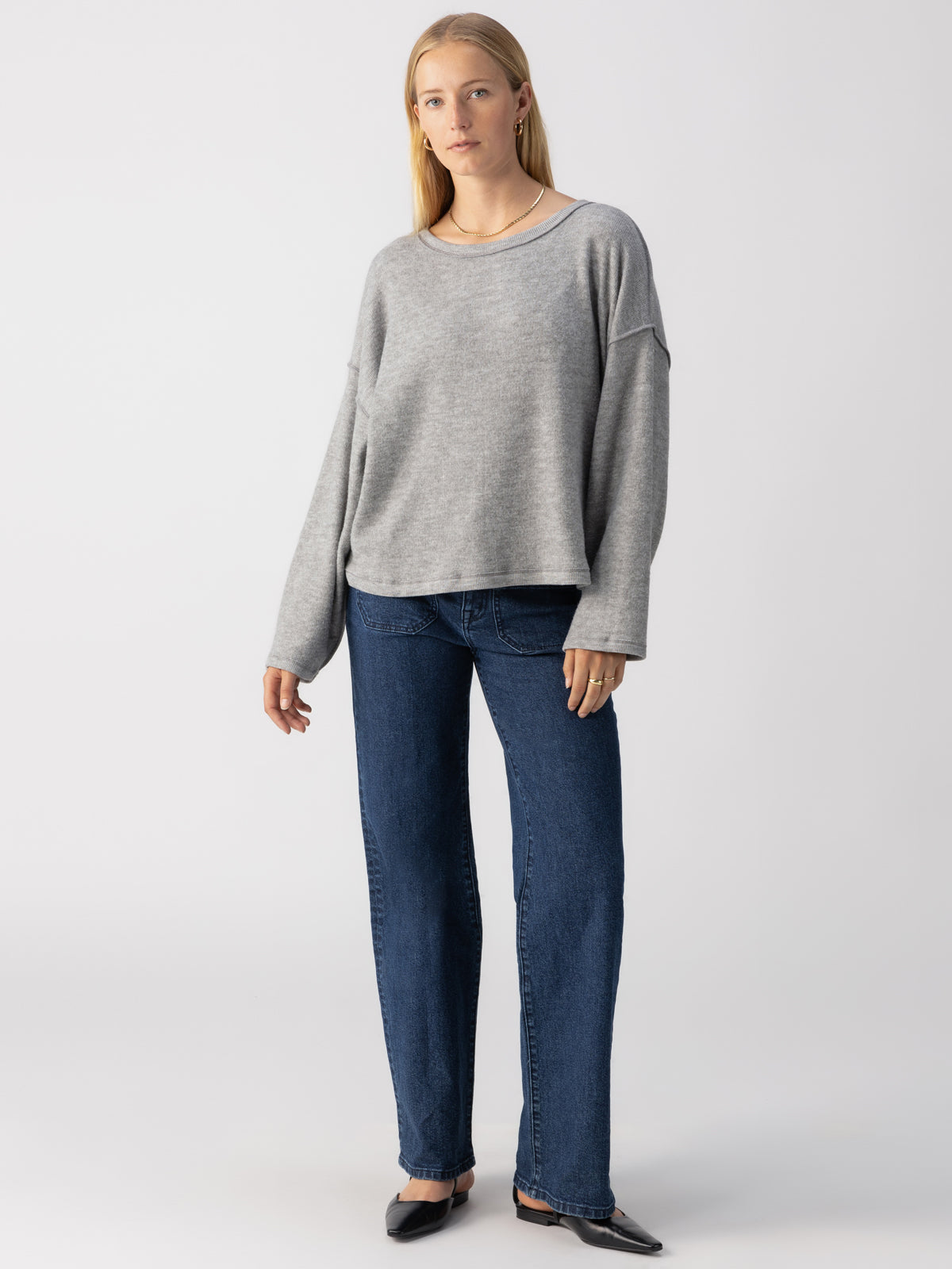 A woman stands against a plain white background wearing the Sanctuary Clothing Change Of Scenery Knit Top in Heather Ash, straight-leg blue jeans, and black pointed shoes. She has long blonde hair and a neutral expression, with her arms relaxed by her sides.