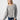 A person with long, straight blonde hair is wearing the Sanctuary Clothing "Change Of Scenery Knit Top" in Heather Ash and blue jeans. They are looking down and slightly to the side, with one arm hanging relaxed by their side against a plain, light gray background.