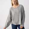 A person with long, straight blonde hair is wearing the Sanctuary Clothing "Change Of Scenery Knit Top" in Heather Ash and blue jeans. They are looking down and slightly to the side, with one arm hanging relaxed by their side against a plain, light gray background.