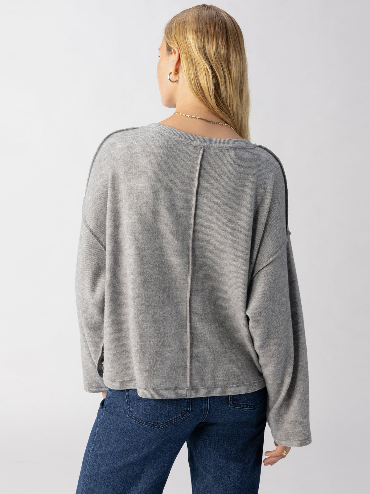 Rear view of a person with long blonde hair wearing the Sanctuary Clothing Change Of Scenery Knit Top in Heather Ash, featuring a visible seam down the center of the back, paired with blue jeans. The person is standing against a plain, light-colored background.