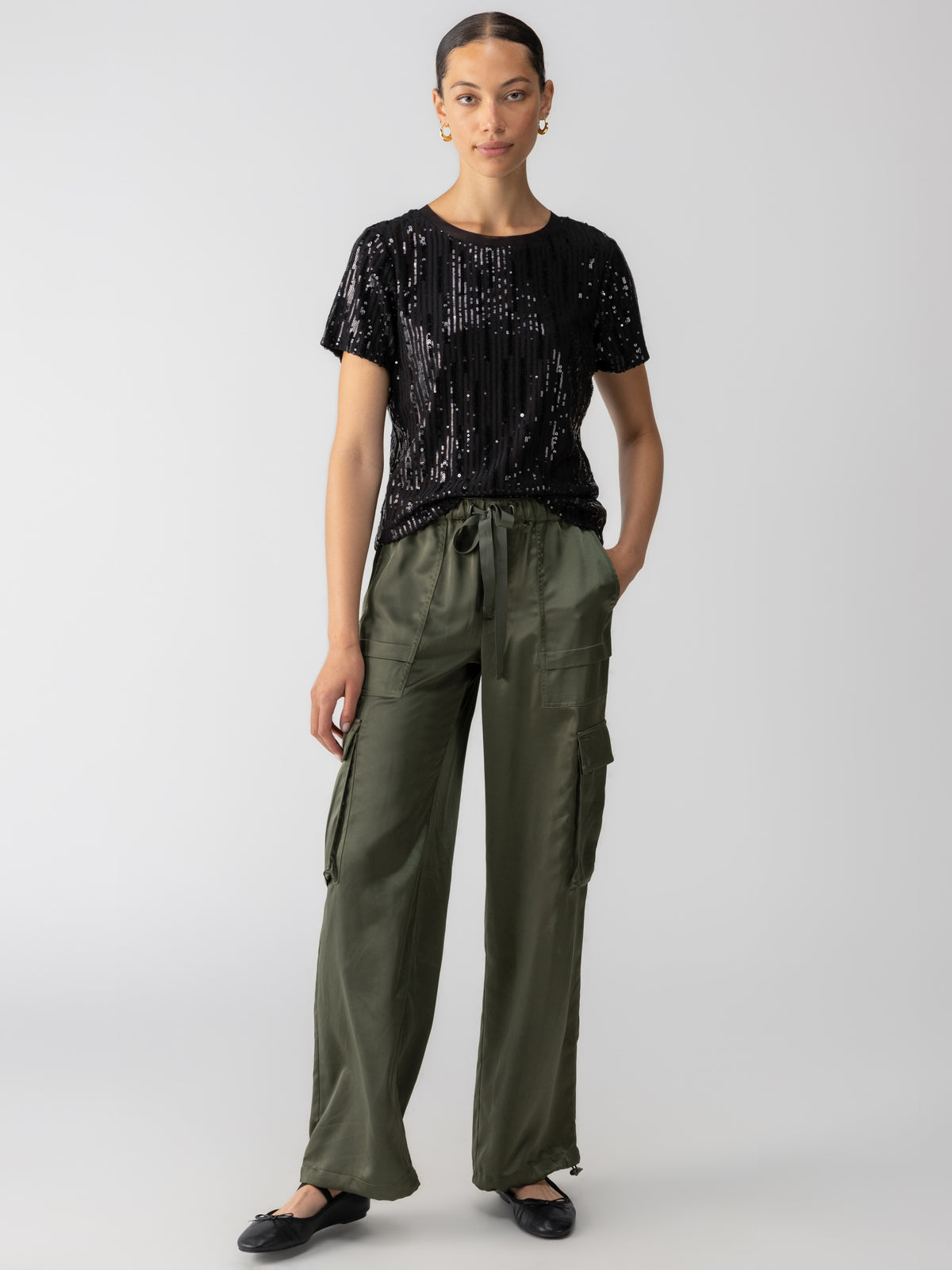 A person is standing against a plain background, wearing the Sequin Perfect Tee Black from Sanctuary Clothing and olive green cargo pants with drawstrings. They have one hand in a pocket of the pants and are wearing black flat shoes. Their hair is pulled back.