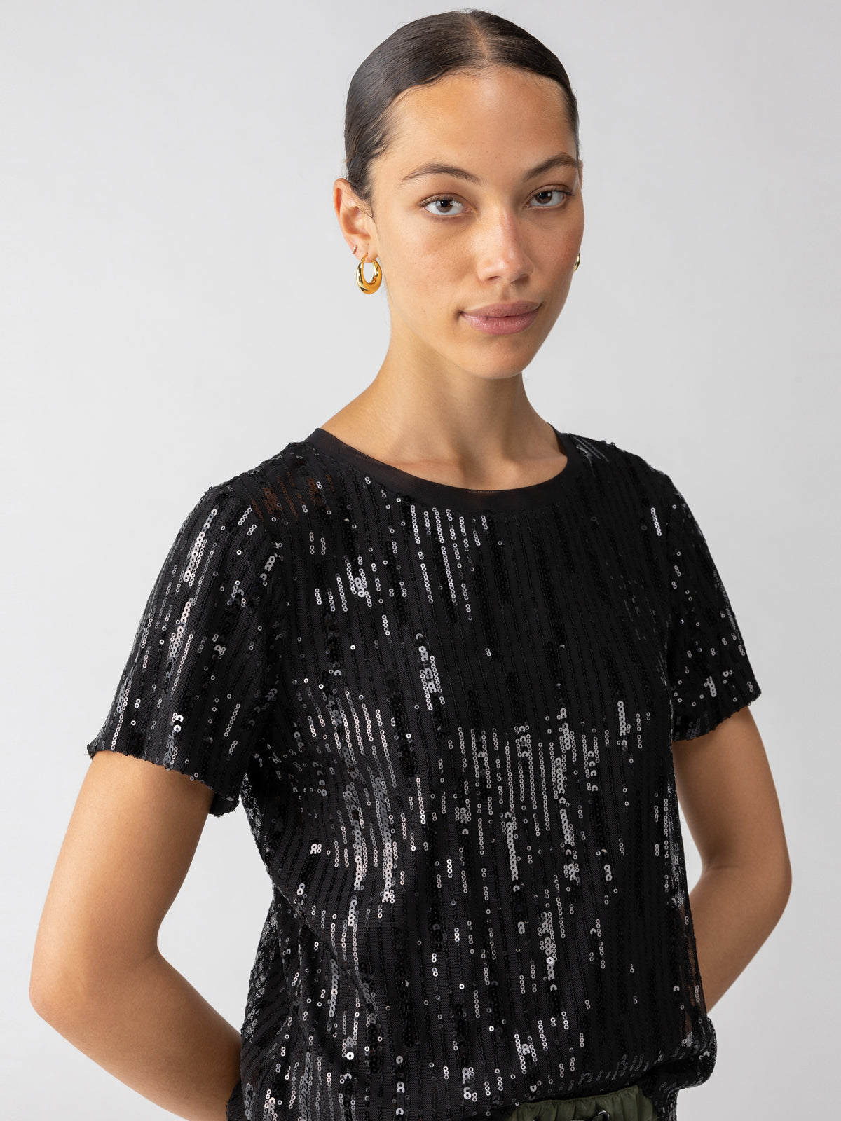 A person with neatly styled, pulled-back hair is wearing the Sequin Perfect Tee in black from Sanctuary Clothing. They have a calm expression and are standing against a plain white background, accessorized with gold hoop earrings.