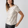 A woman with long brown hair is wearing the Sanctuary Clothing 'Sequin Perfect Tee Lets Celebrate,' a short-sleeved, beige top adorned with silver sequins, paired with black pants. She is posing with one hand resting on her hip, looking confidently at the camera against a plain gray background.