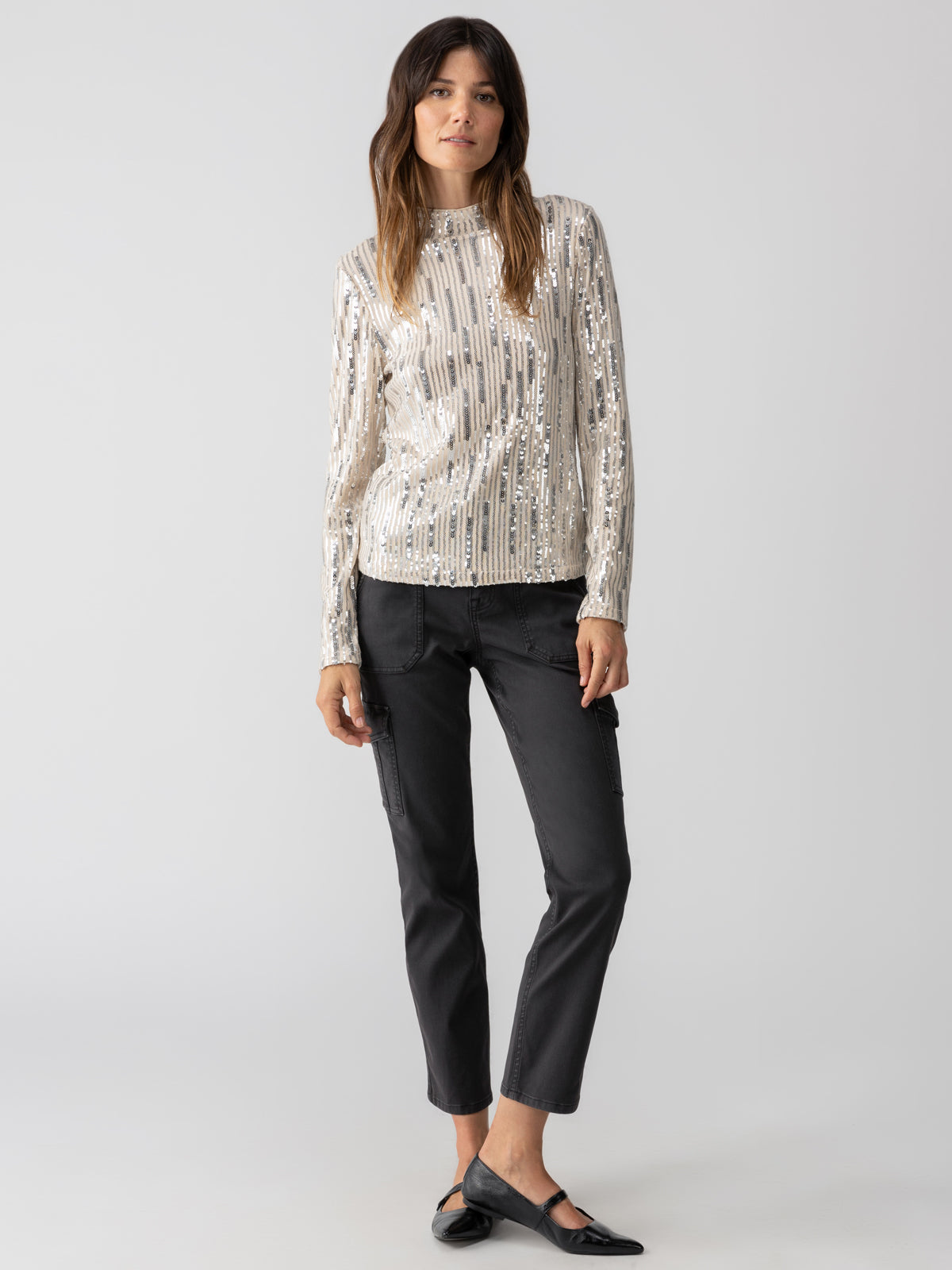 A person with long hair stands against a plain background, wearing the Sequin Mock Top Lets Celebrate from Sanctuary Clothing, paired with dark gray pants and black flat shoes. They have a relaxed expression and their arms hang naturally by their sides.