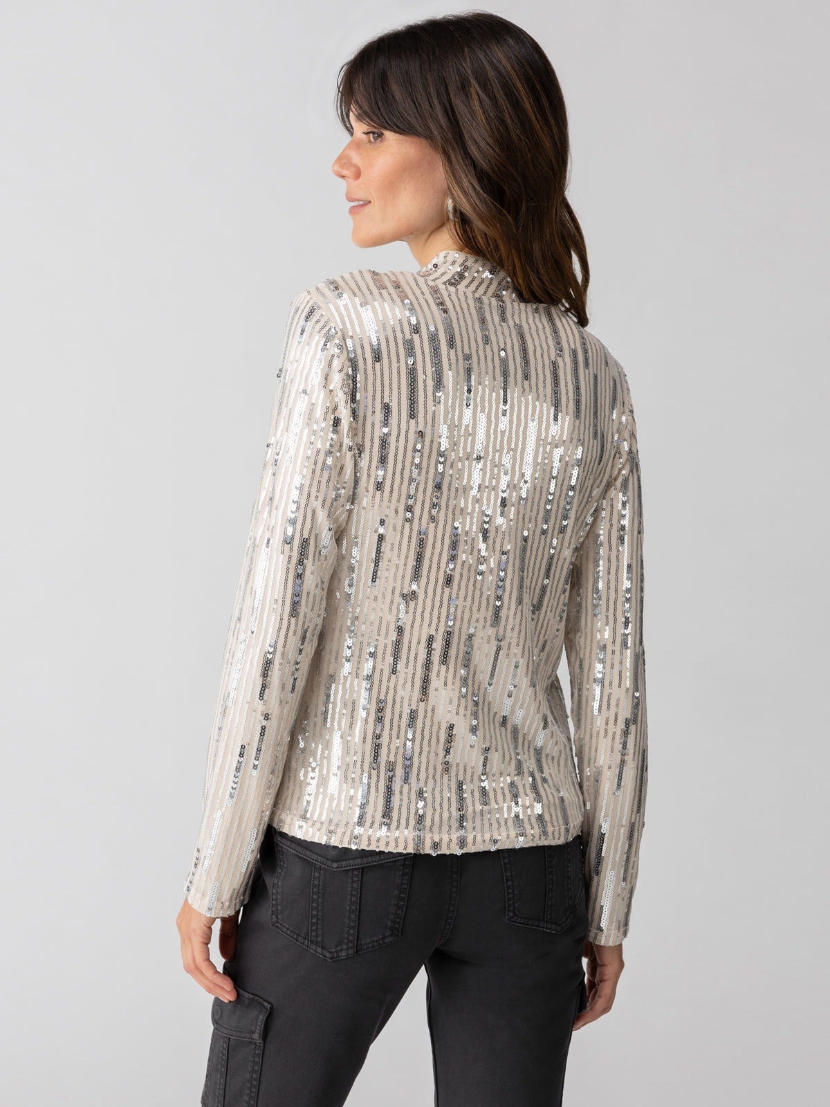 A woman with shoulder-length hair wearing Sanctuary Clothing's shiny, silver "Sequin Mock Top Lets Celebrate" stands facing away, slightly turned to the side. She is also wearing dark pants, and the background is plain and light-colored.