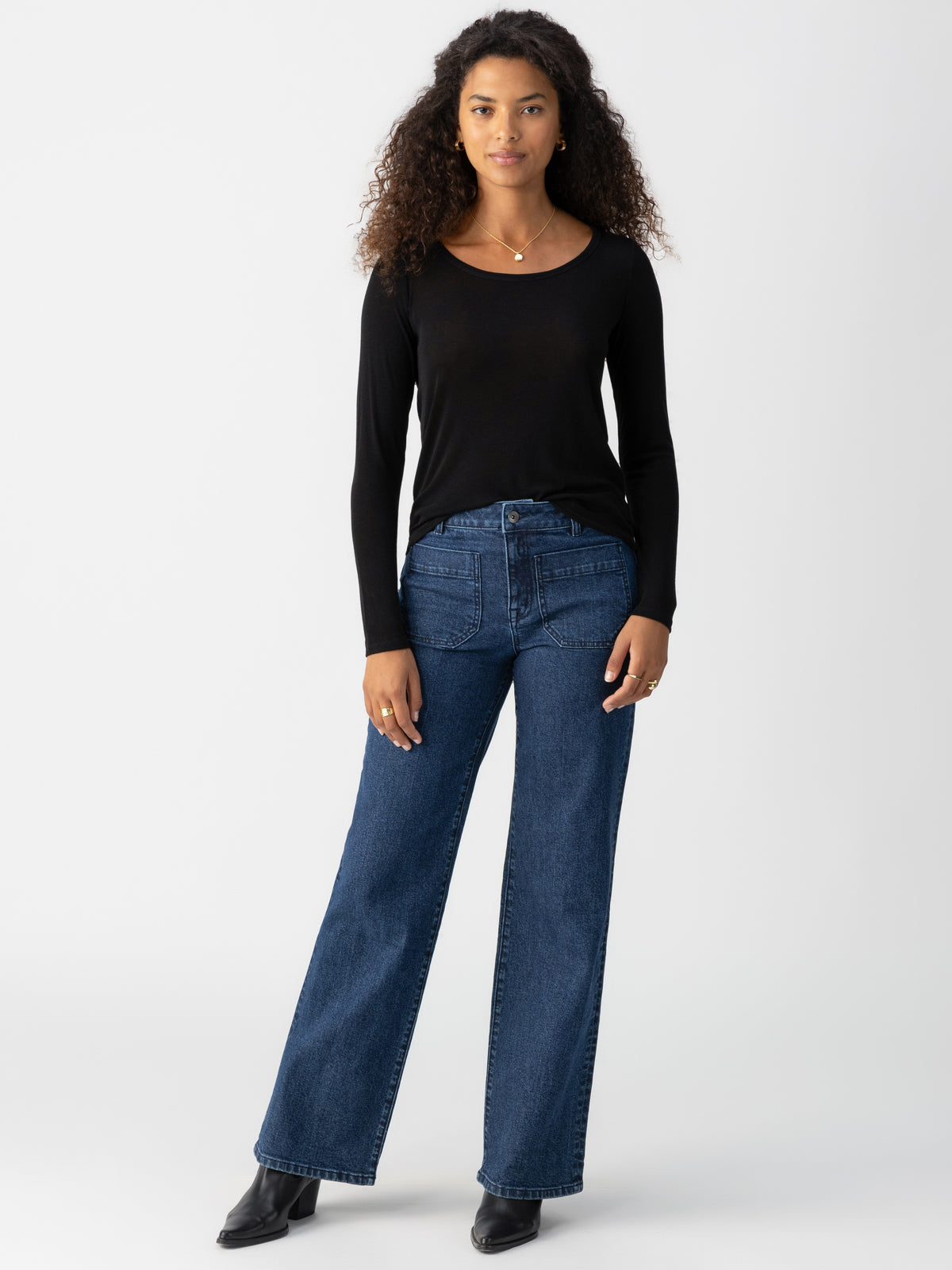 A woman with long curly hair is wearing Sanctuary Clothing's "Keep It Simple Tee" in black, paired with high-waisted blue wide-leg jeans. She completes her outfit with black boots and a simple necklace, standing against a plain white background.