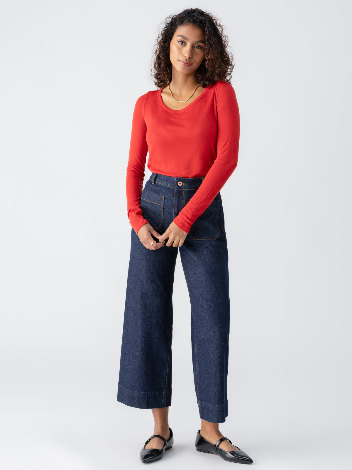 A person with curly hair stands against a plain background, wearing the Sanctuary Clothing Keep It Simple Tee in Cherry Red and high-waisted, wide-leg blue jeans. They have on black flat shoes and are clasping their hands in front of them, with a relaxed expression.