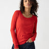 A person with curly hair is wearing a long-sleeved Keep It Simple Tee in Cherry Red from Sanctuary Clothing paired with dark jeans. They are posing against a light gray background with one hand in their pocket and a subtle smile, accessorized with a thin gold necklace.