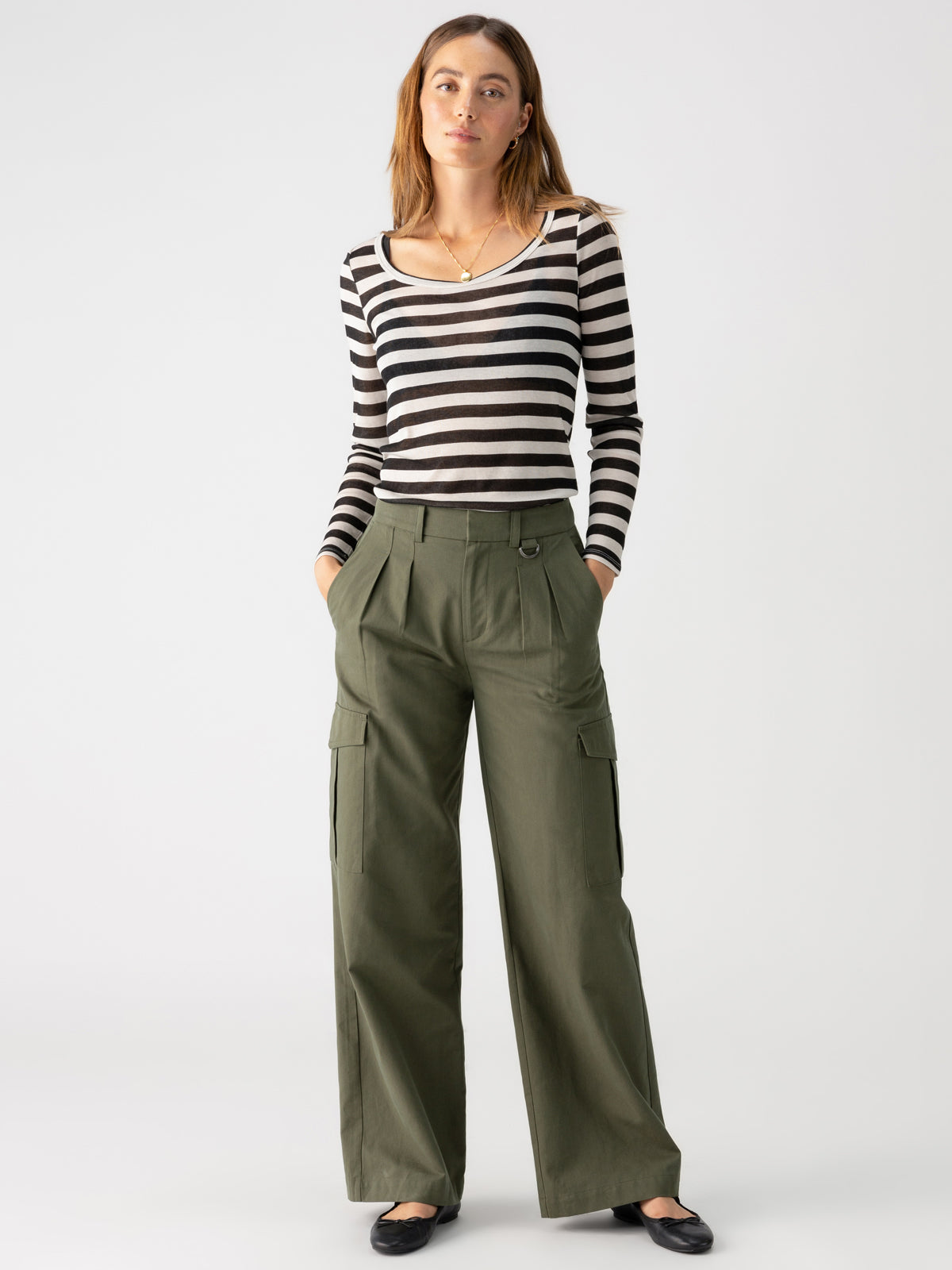 A person with long, light brown hair wears the Keep It Simple Tee Toasted Almond Black Stripe from Sanctuary Clothing paired with olive green high-waisted, wide-leg cargo pants. They have their hands in their pockets and are wearing black shoes, posing against a plain white background.