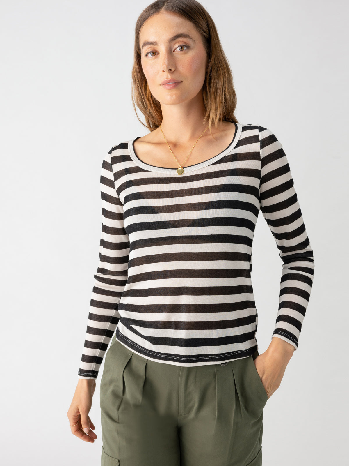 A person with shoulder-length brown hair is standing against a plain white background. They are wearing the Keep It Simple Tee Toasted Almond Black Stripe by Sanctuary Clothing, paired with olive green pants. They have a neutral expression and one hand in their pocket.