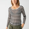 A person with shoulder-length brown hair is standing against a plain white background. They are wearing the Keep It Simple Tee Toasted Almond Black Stripe by Sanctuary Clothing, paired with olive green pants. They have a neutral expression and one hand in their pocket.