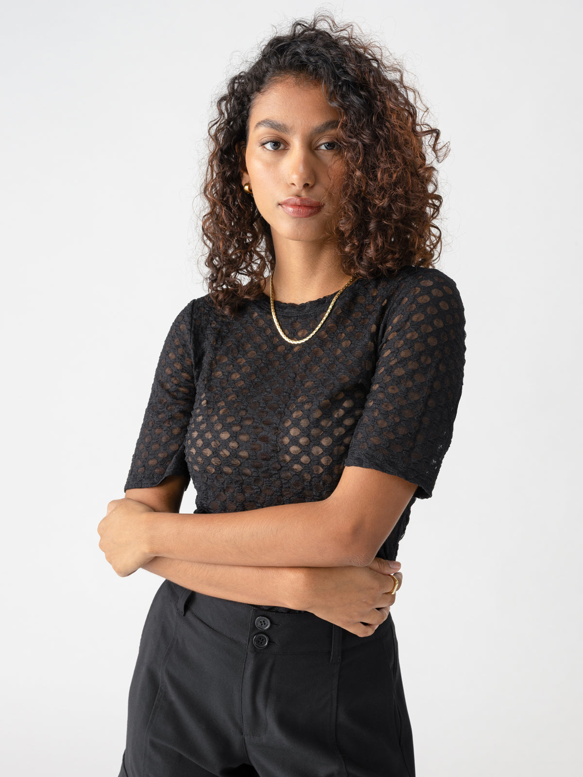 A person with curly hair stands against a plain background, wearing Sanctuary Clothing's short-sleeved Geo Mesh Tee in black and black pants, accessorized with a gold necklace. Their arms are crossed, and they have a neutral expression.