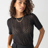 A person with curly hair poses confidently, wearing the Sanctuary Clothing Geo Mesh Tee Black over a dark bra, paired with high-waisted black pants and a gold chain necklace. The person has their hands in their pockets, standing against a light grey background.