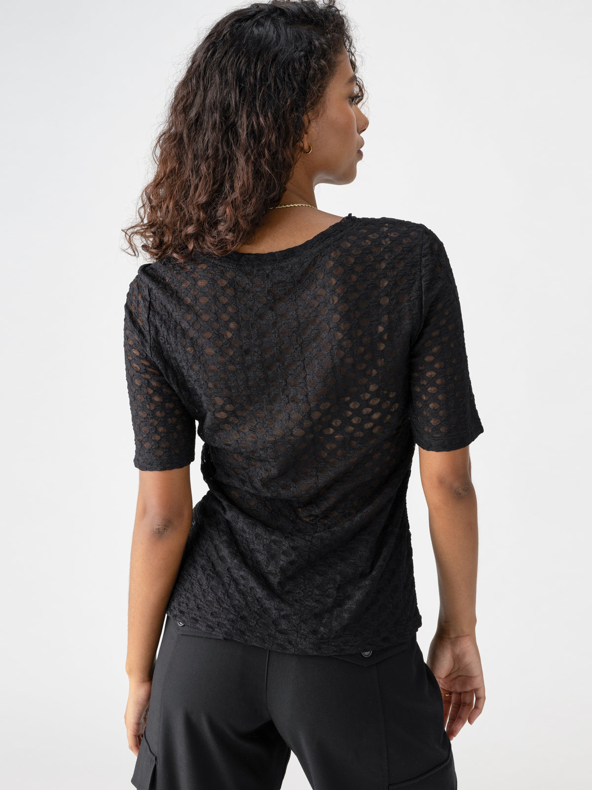 A person with curly hair is standing with their back to the camera, wearing the Sanctuary Clothing Geo Mesh Tee Black and black pants. The background is plain and light-colored.