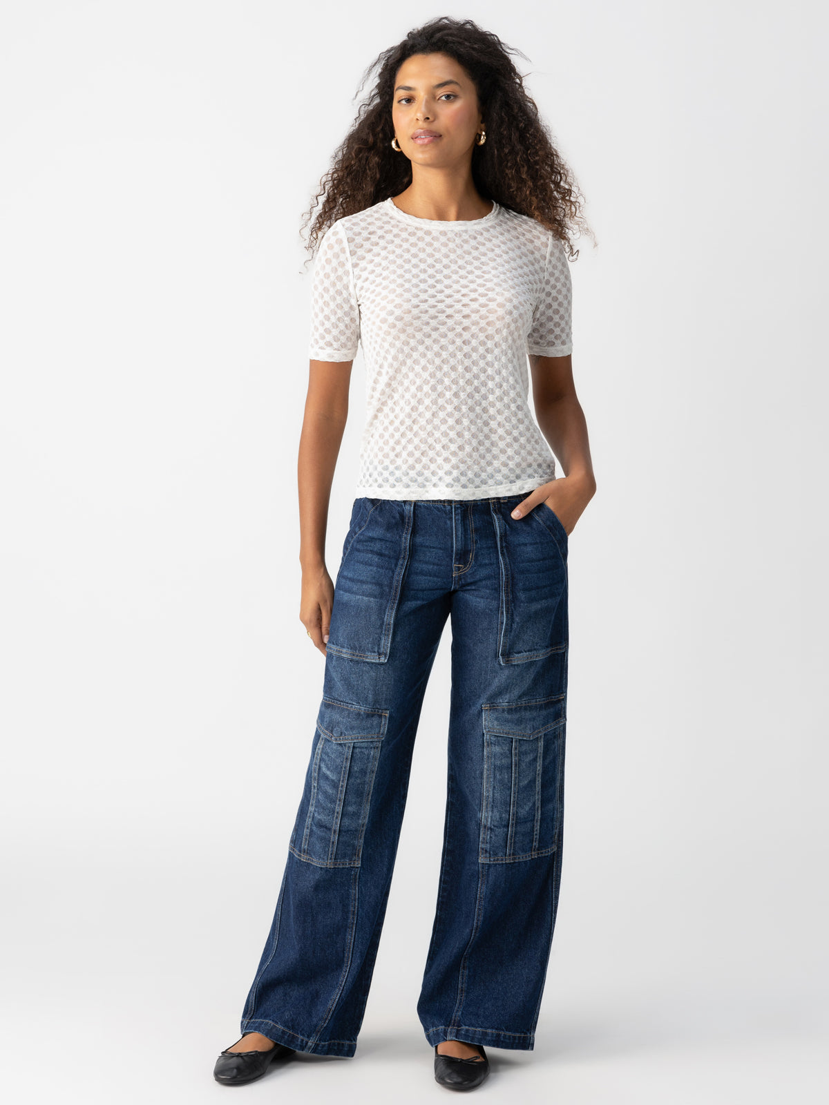 A woman with curly hair wearing the Geo Mesh Tee Chalk from Sanctuary Clothing and dark blue wide-leg jeans with multiple pockets stands against a plain background. She is looking forward with one hand in her pocket and the other arm relaxed by her side.