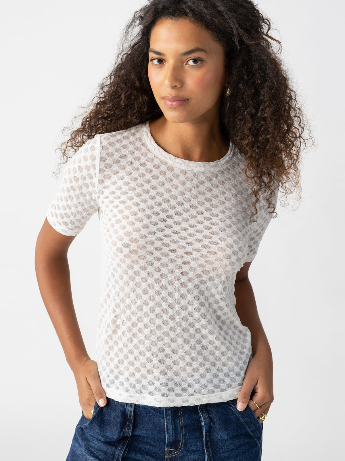 A person with long curly hair is wearing the Geo Mesh Tee Chalk from Sanctuary Clothing, a white, semi-sheer, short-sleeve top featuring a dotted pattern. They have their hands in the pockets of their blue jeans and are standing against a plain white background, looking directly at the camera with a neutral expression.