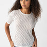 A person with long curly hair is wearing the Geo Mesh Tee Chalk from Sanctuary Clothing, a white, semi-sheer, short-sleeve top featuring a dotted pattern. They have their hands in the pockets of their blue jeans and are standing against a plain white background, looking directly at the camera with a neutral expression.