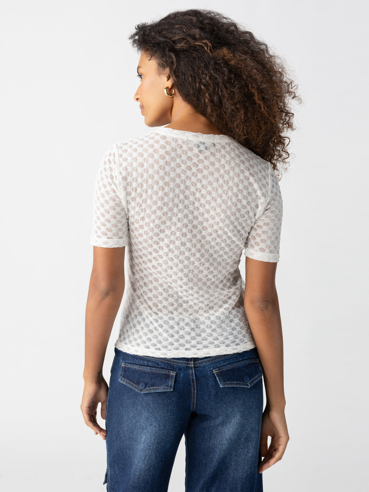 A person with curly hair is shown from the back, wearing the Geo Mesh Tee Chalk by Sanctuary Clothing and blue jeans. They are looking to the side, with one arm slightly bent, against a plain white background.