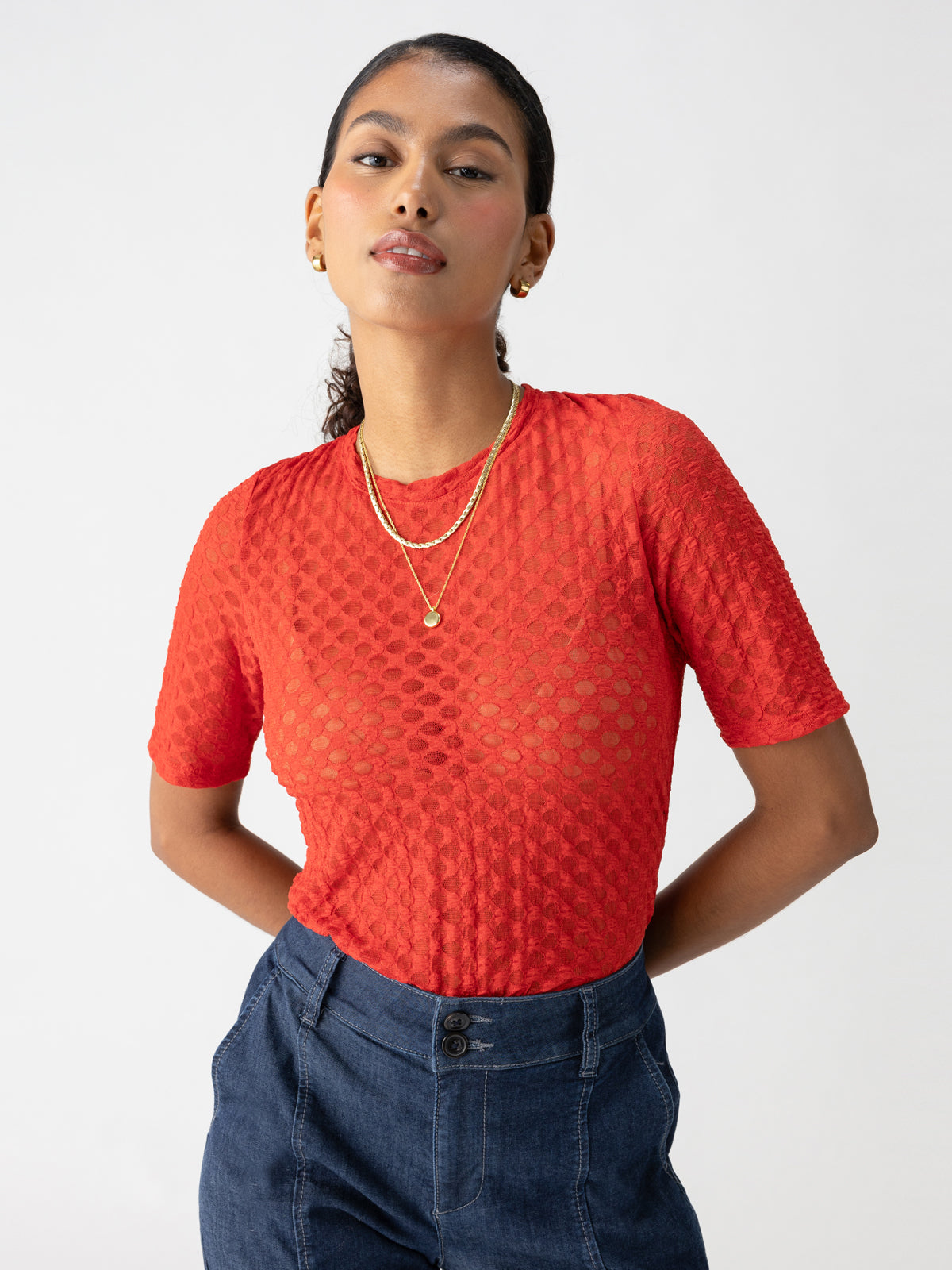 A person with long dark hair tied back is wearing the Sanctuary Clothing Geo Mesh Tee in Cherry Red over a sheer undershirt. They have gold hoop earrings and a layered gold necklace. They are also wearing high-waisted blue pants and posing with their hands behind their back.