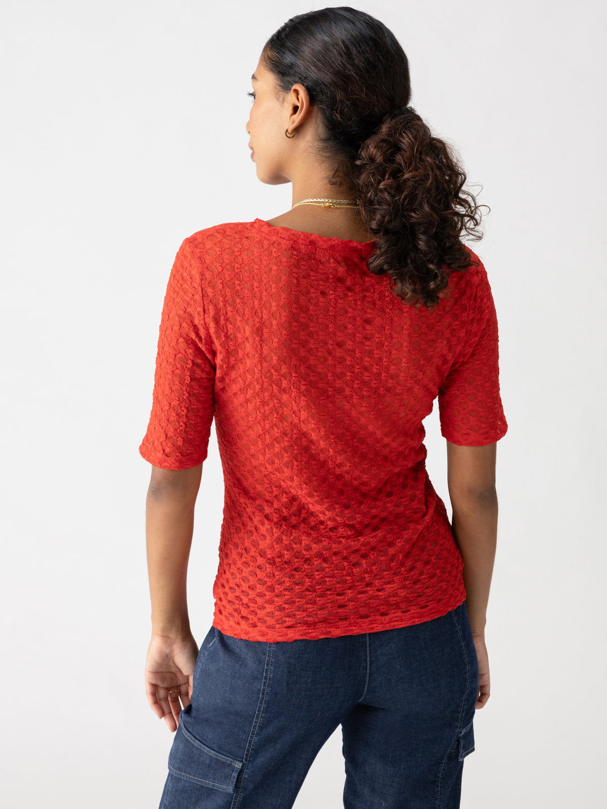 A person with dark curly hair is seen from the back, wearing the Geo Mesh Tee Cherry Red by Sanctuary Clothing, styled with elbow-length sleeves and paired with dark blue denim pants. The individual has a simple gold necklace, and their hair is tied in a low ponytail. The background features a light, neutral color.