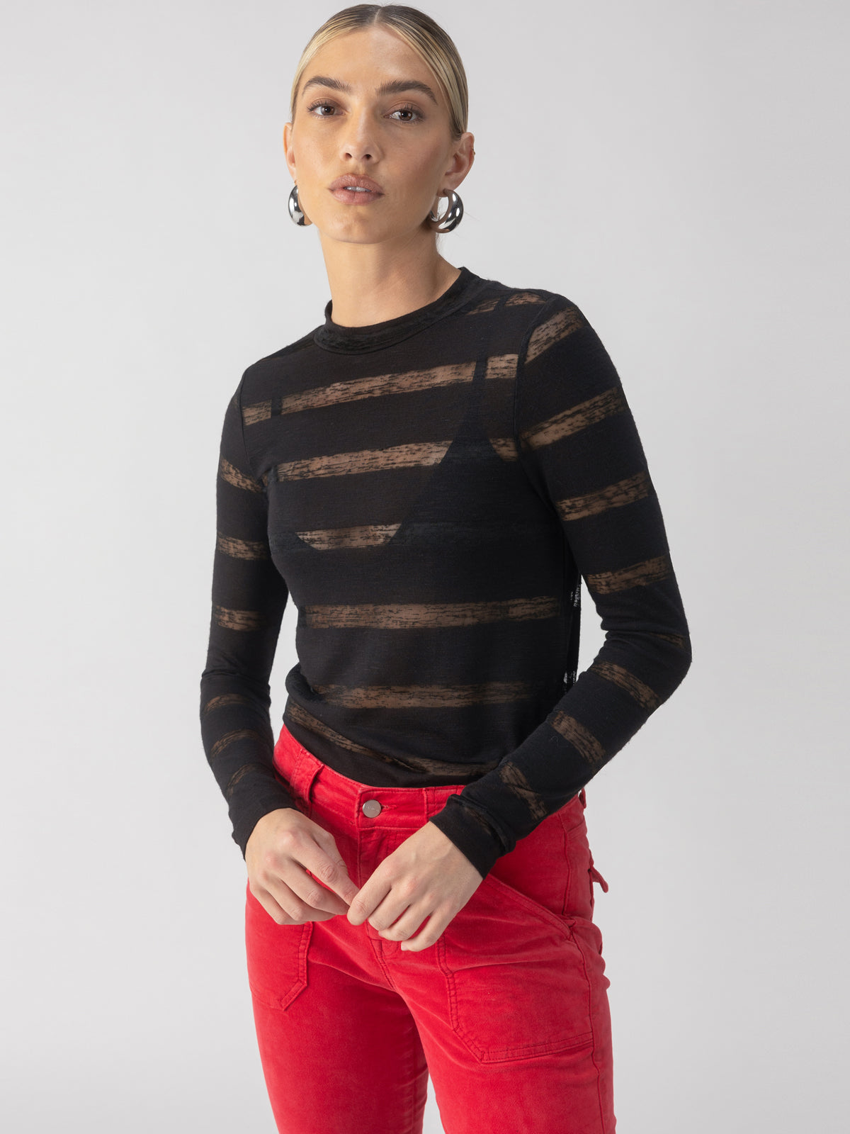 A person with short blonde hair wears the Sanctuary Clothing's Semi Sheer Stripe Tee in black paired with vibrant red pants. They stand against a neutral background, hands resting on their hips.