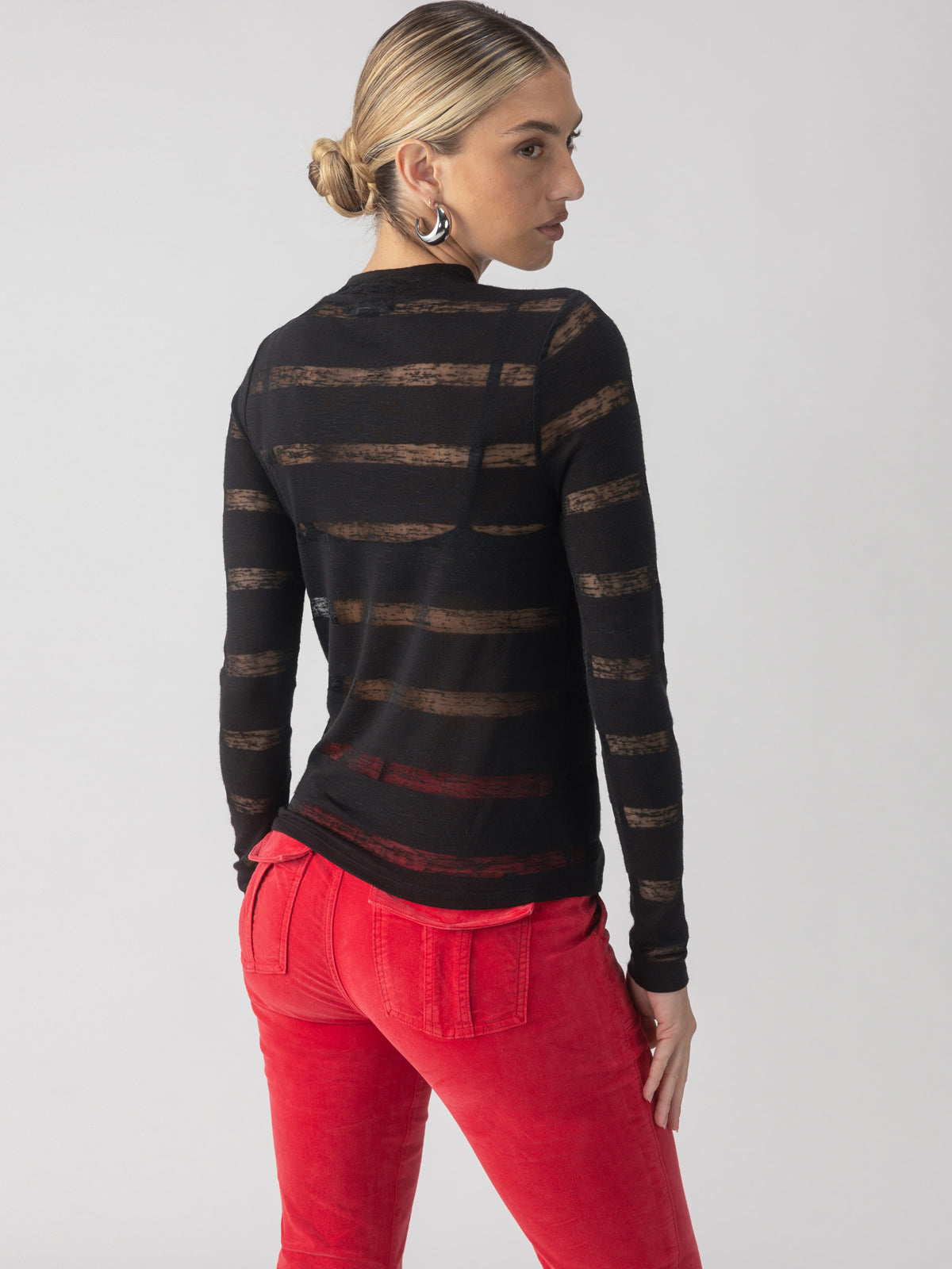 A person with blonde hair tied back is wearing the Sanctuary Clothing Semi Sheer Stripe Tee in black, paired with bright red pants. They are standing and looking over their left shoulder against a plain background.