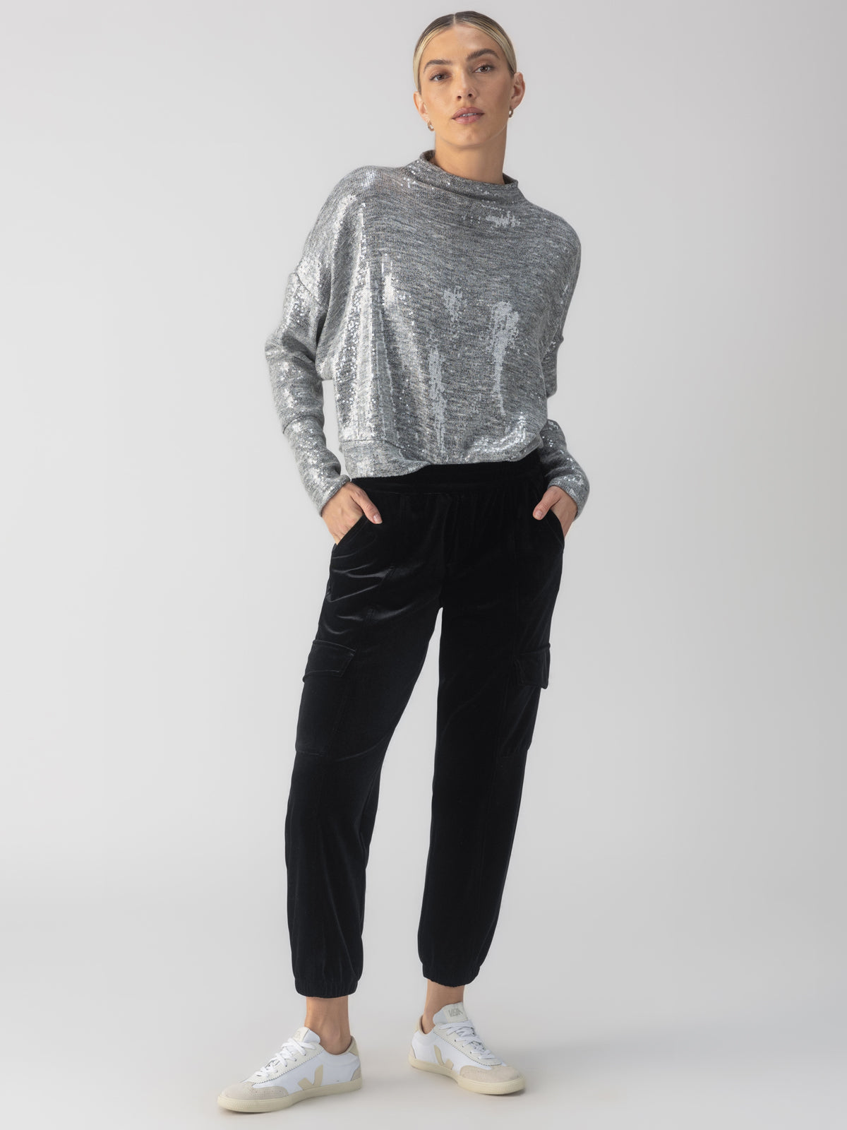 A person stands against a neutral background wearing the Sequin Funnel Neck Top in Heather Grey by Sanctuary Clothing, paired with black velvet jogger pants and white sneakers. They have short hair, styled back neatly.