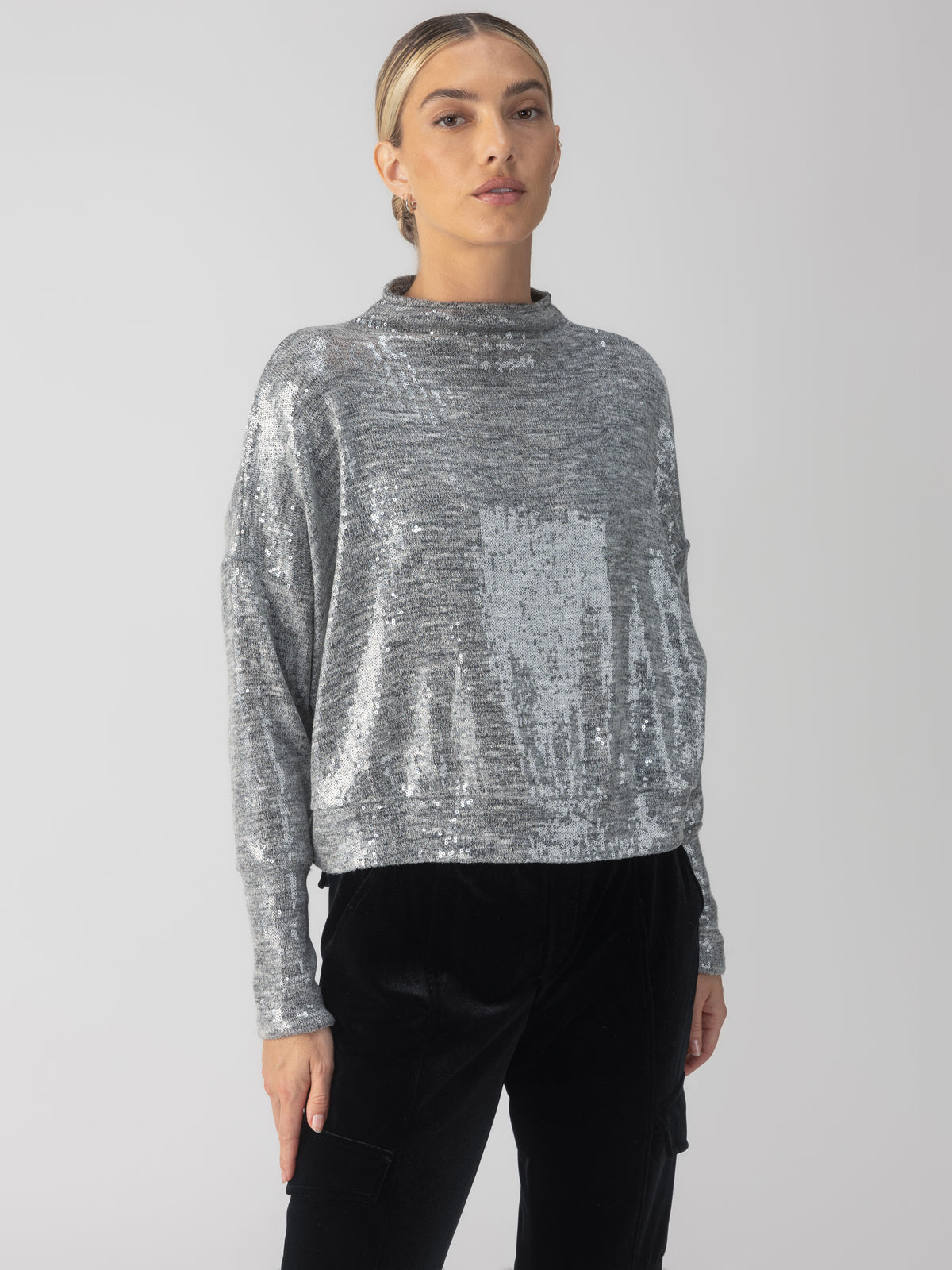 A person wearing the Sequin Funnel Neck Top in Heather Grey from Sanctuary Clothing pairs it with black pants while standing against a plain background. The funnel neck top features a shimmering texture and a relaxed fit. The person's hair is styled neatly, pulled back for an elegant look.