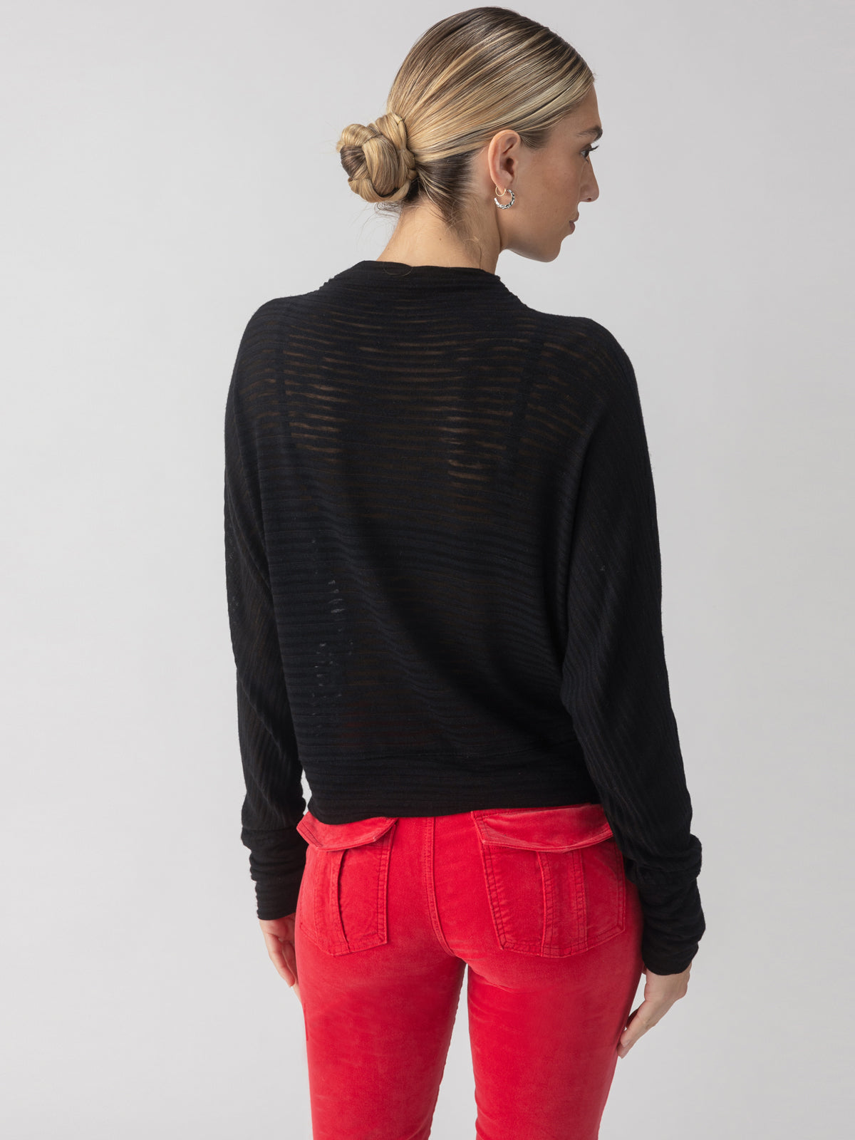 A person with blonde hair styled in a bun is wearing the Sanctuary Clothing Sheer Stripe Funnel Neck Top in black, paired with red pants. They are standing with their back to the camera against a plain background.