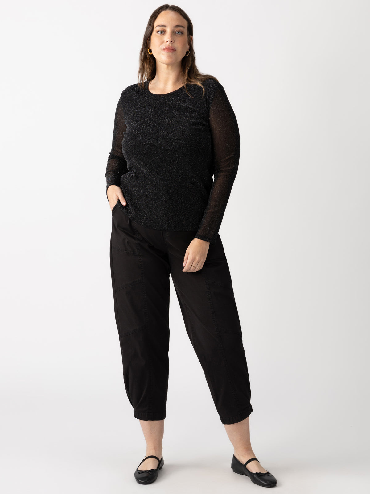 A woman stands against a white background wearing the Sahara Tapered Pant Black from Sanctuary Clothing's Inclusive Collection. She has long hair and wears black flats, with her hands casually resting in her pant pockets as she looks forward.