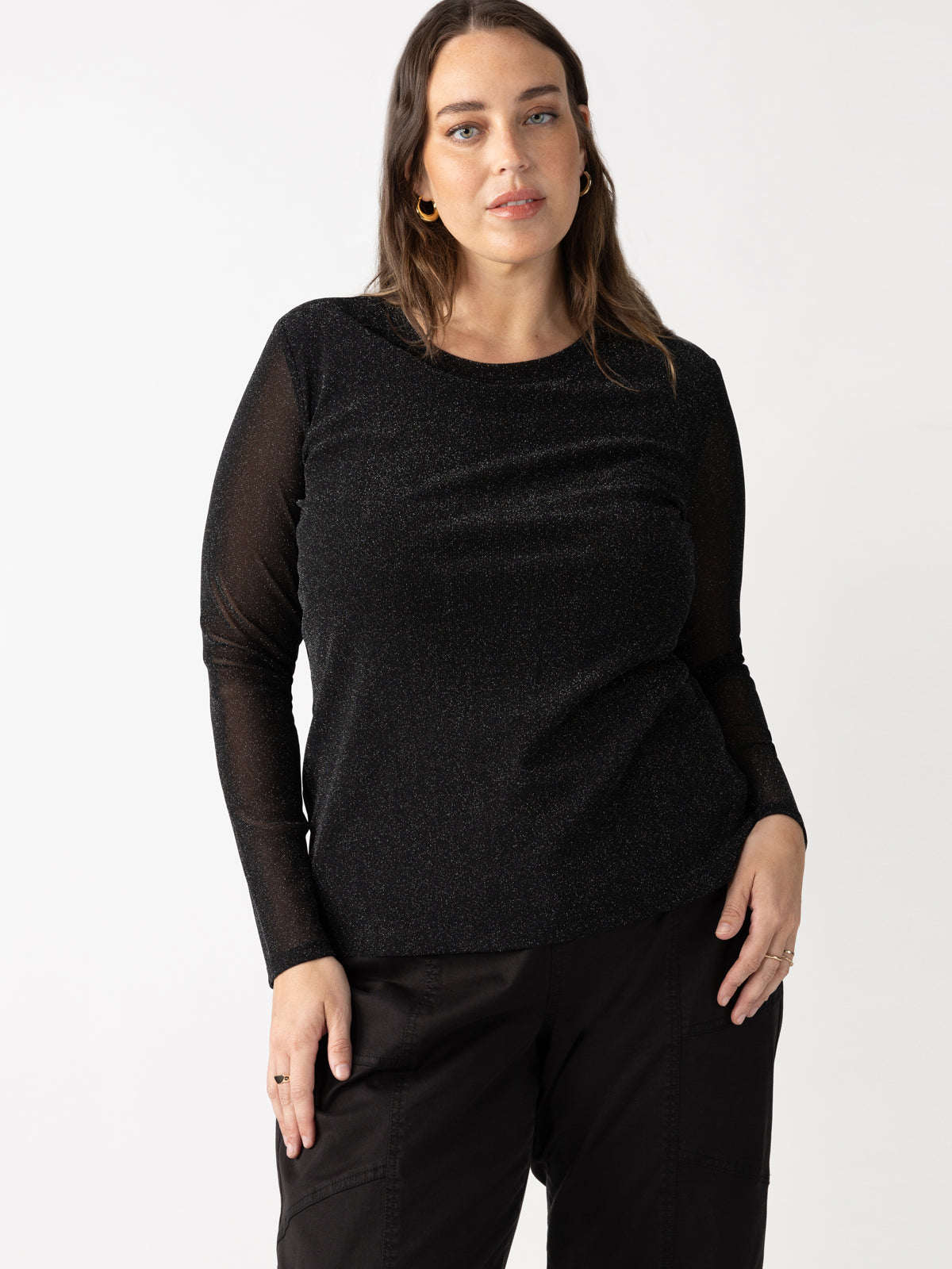 A woman with long brown hair is wearing the Sparkle Mesh Perfect Tee Black from Sanctuary Clothing's Plus Size Collection and dark pants. She is standing against a plain white background with her left hand resting on her hip, displaying a calm expression.