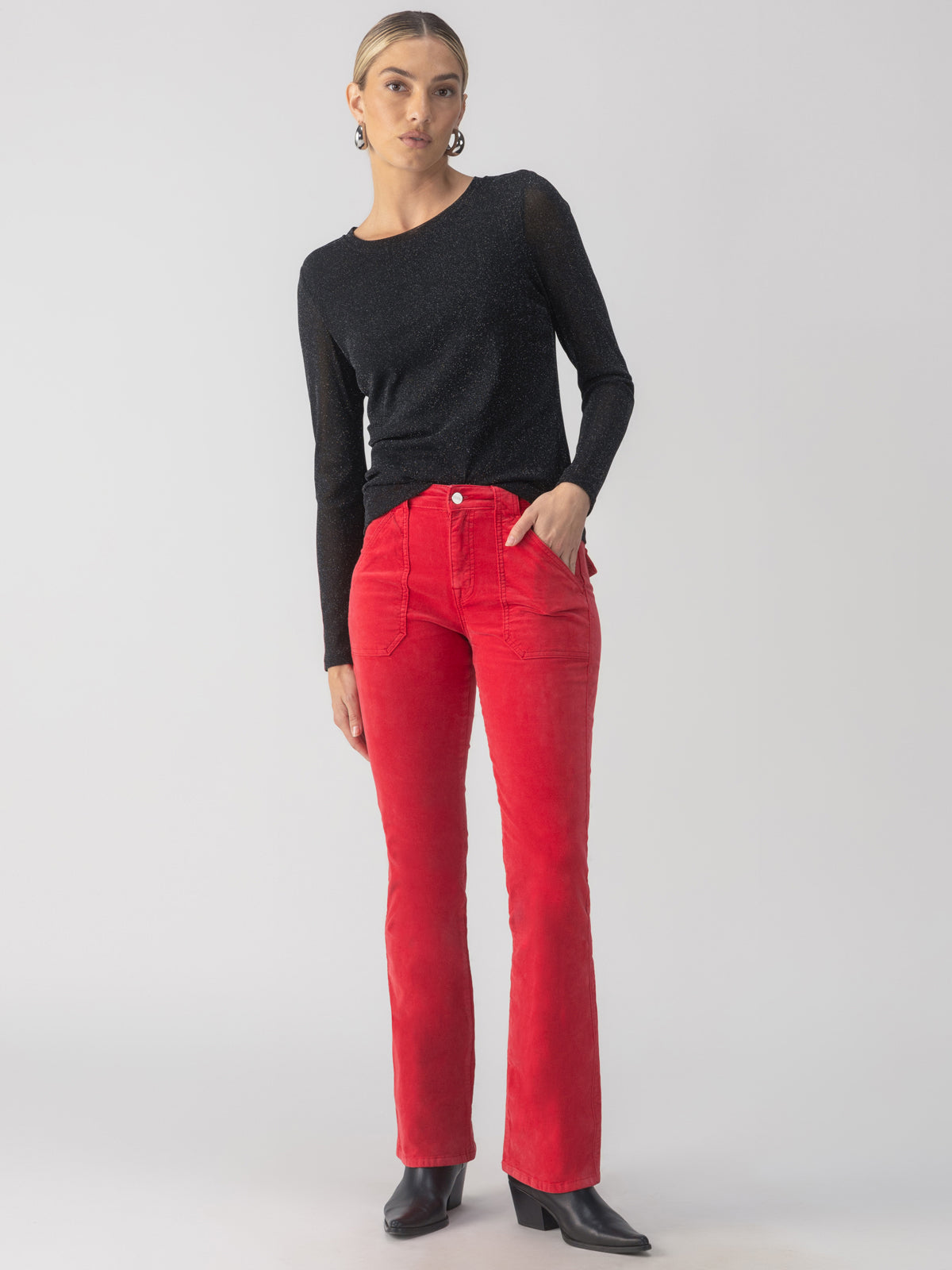 A person with blonde hair stands against a plain background, wearing the Sanctuary Clothing's sparkle mesh perfect tee in black and bright red pants. They are also wearing black ankle boots. Their hands are in their pockets, and they have a neutral expression.