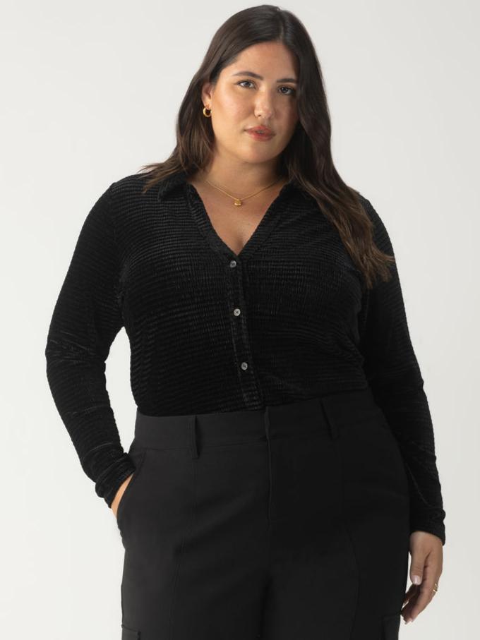 A woman with long, dark hair is confidently wearing a textured velvet button-up top from the Sanctuary Clothing black plus size collection. She stands against a plain background, looking directly at the camera.