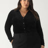 A woman with long, dark hair is confidently wearing a textured velvet button-up top from the Sanctuary Clothing black plus size collection. She stands against a plain background, looking directly at the camera.