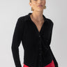 A woman stands confidently wearing a textured velvet button-up top in black from Sanctuary Clothing, paired with red pants. Her hair is pulled back, and she accessorizes with large round earrings against a plain, light-colored background.