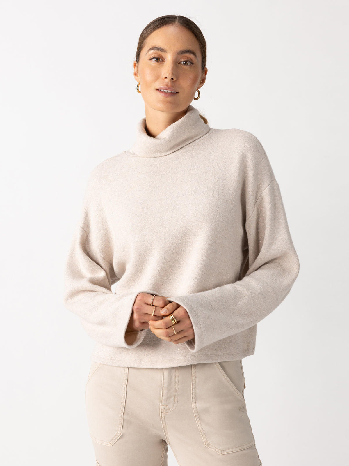 A person dressed in the Cozy Lurex Popover Top Coconut Smoke from Sanctuary Clothing and matching beige pants stands against a plain white background. Their long hair is pulled back, and they’re holding their hands together in front. They adorn hoop earrings and several rings.