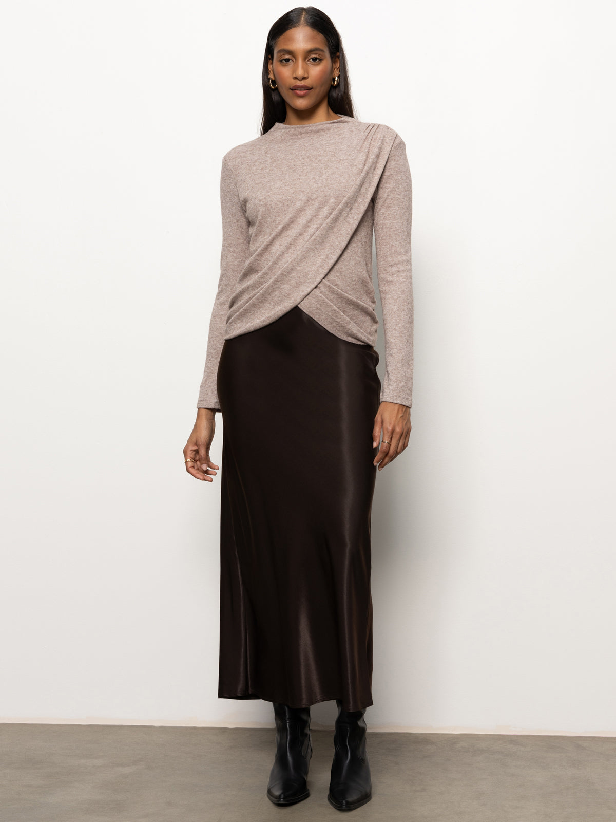 A woman stands against a plain background, wearing the Long Sleeve Mock Draped Top in Heather Cocoa by Sanctuary Clothing along with a long, dark brown skirt. She completes her outfit with black ankle boots and small hoop earrings. Her hair is down, and she has a neutral expression.