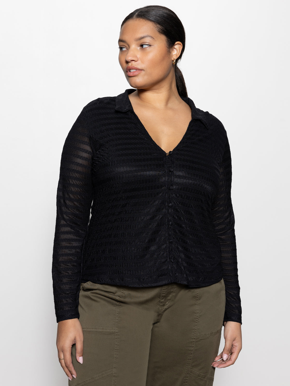 An individual wearing Sanctuary Clothing's mesh lace shirt in black from their inclusive collection, paired with olive green pants, stands against a plain white background. Their dark hair is tied back and they are gazing to the side.