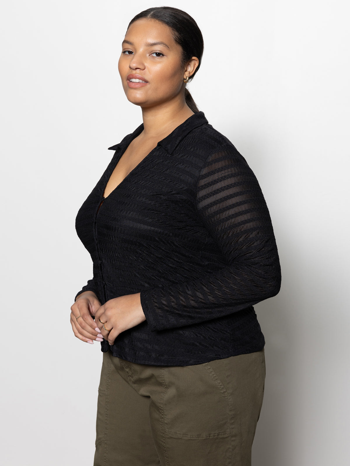 A woman, dressed in the Sanctuary Clothing mesh lace shirt from the inclusive collection, paired with olive green pants, stands against a plain light background. Her hair is pulled back as she confidently gazes to the side.