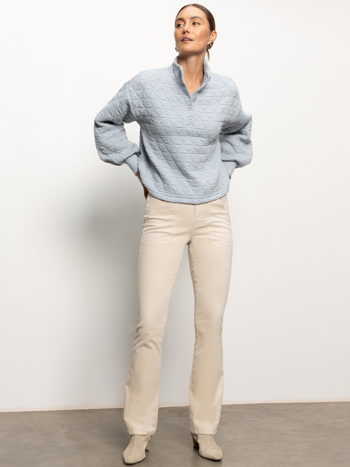 A person stands confidently against a plain background, donning the Quilted Button Up Popover Top in Pearl Blue by Sanctuary Clothing, paired with beige pants and light-colored shoes. Their hands rest on their hips as they gaze slightly upward.