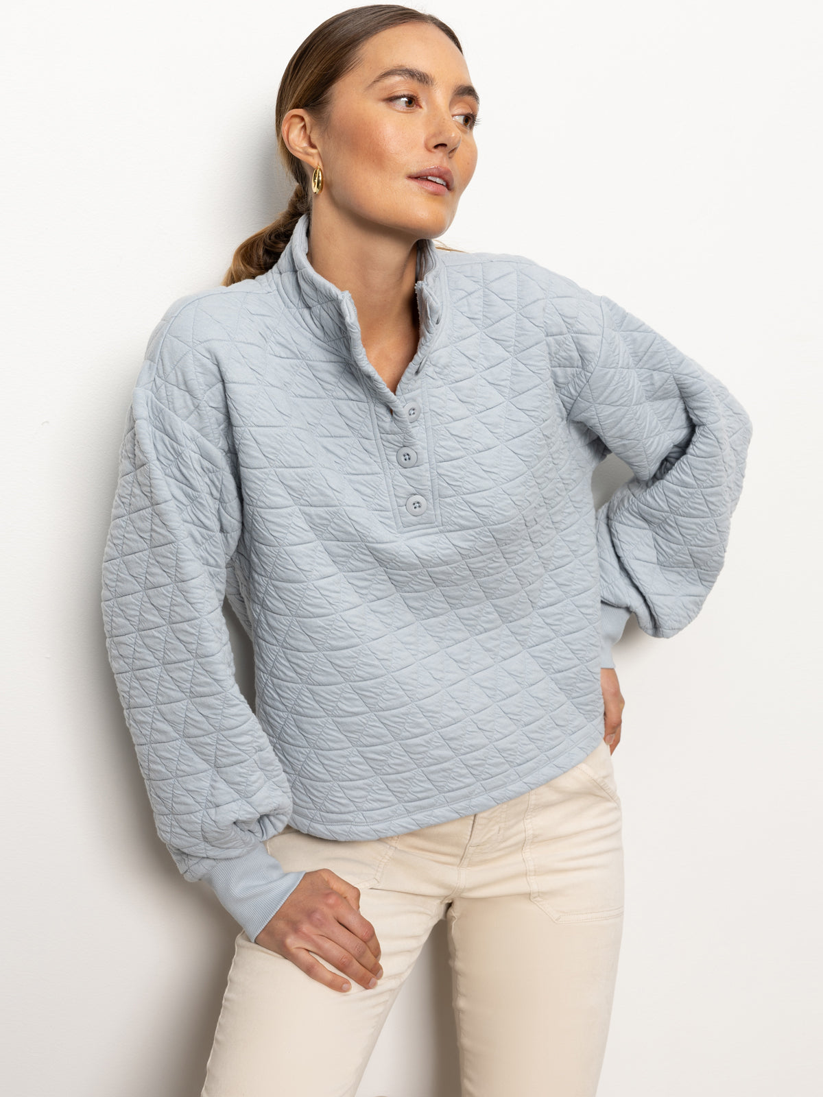 A person wearing Sanctuary Clothing's Quilted Button Up Popover Top in Pearl Blue, paired with off-white pants. They are leaning against a white wall with their hands in their pockets.