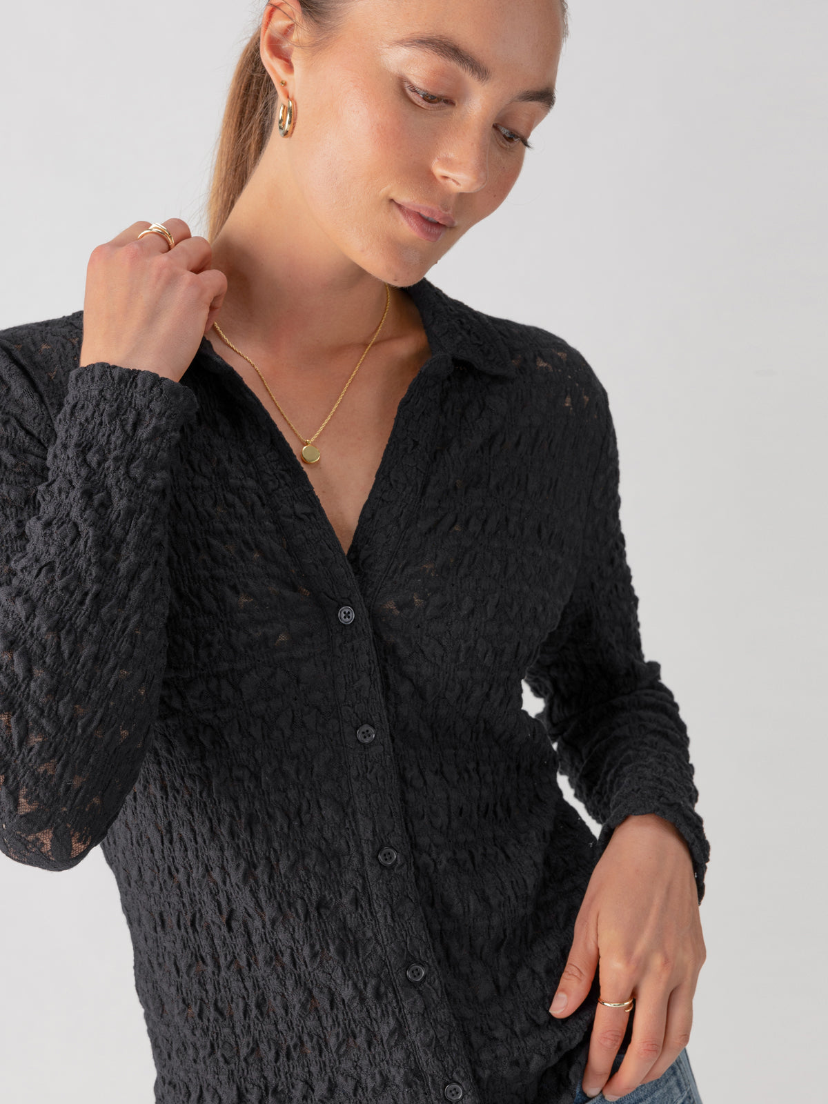 A person is wearing the Dreamgirl Lace Button Up Black blouse from Sanctuary Clothing, paired with light blue jeans and gold jewelry, including hoop earrings, a necklace, and rings. They have light brown hair styled back and are looking down with a hand touching the collar.