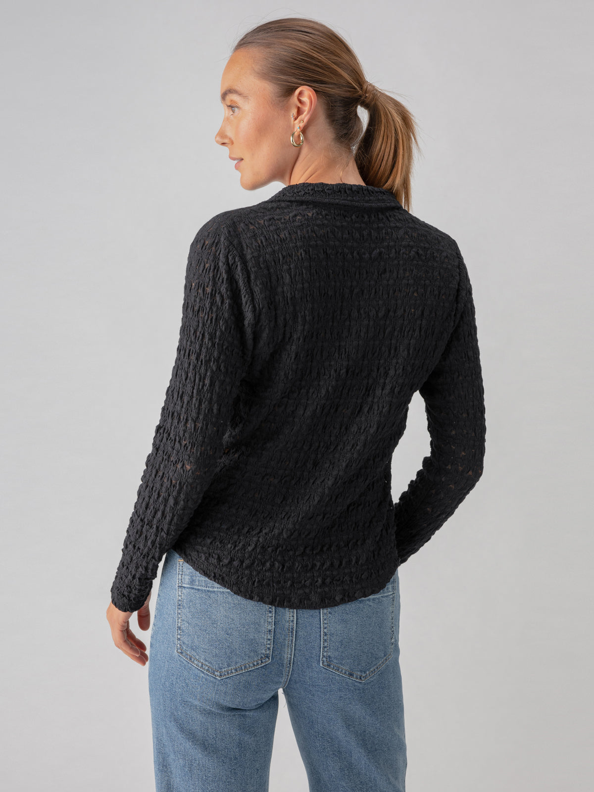 A person with light hair tied back in a ponytail is seen from behind, wearing the Dreamgirl Lace Button Up Black by Sanctuary Clothing along with light blue jeans. The background is plain and light-colored, providing a simple contrast to the dark top.