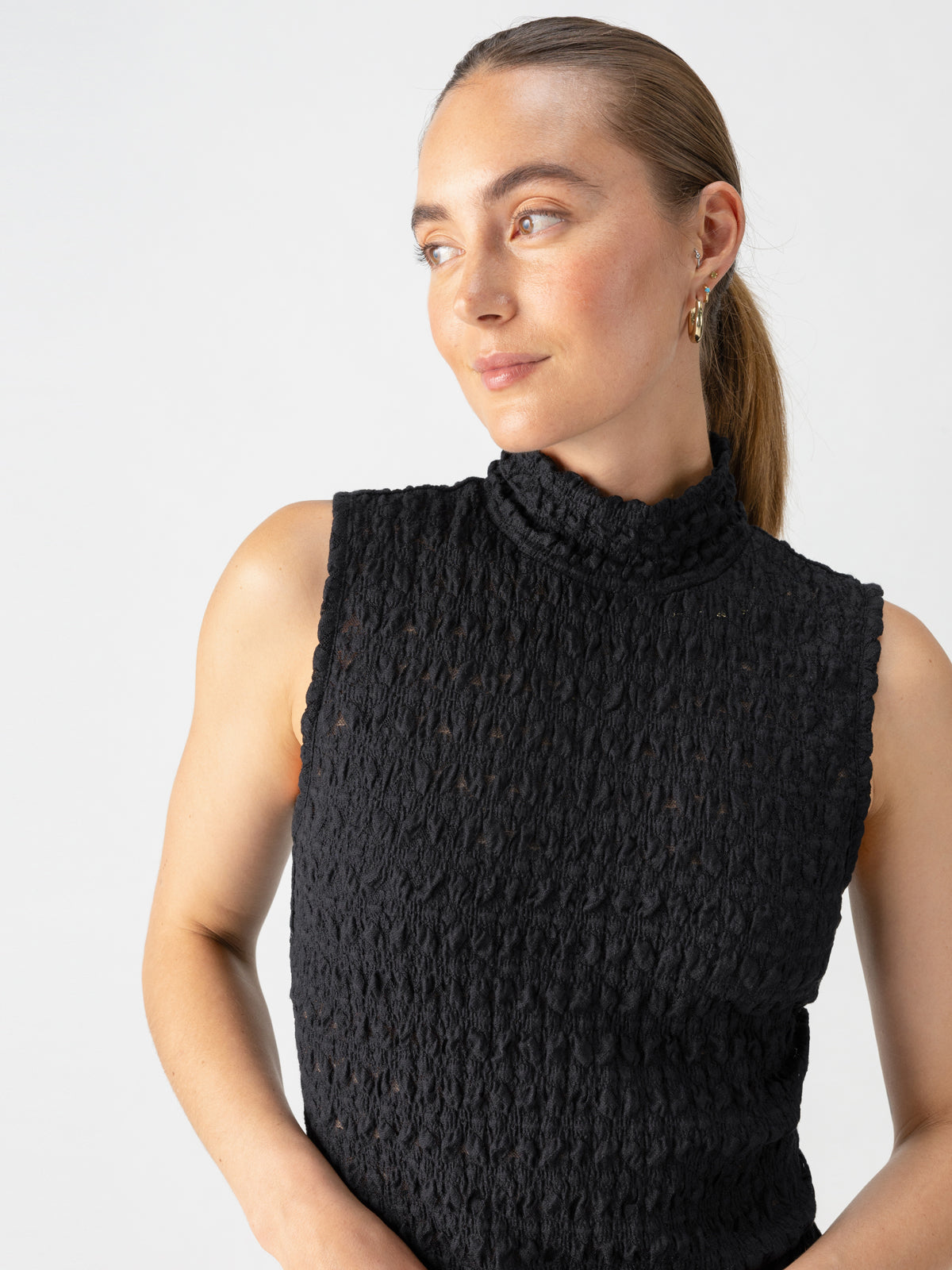 A person with light skin and light brown hair pulled back is wearing the Pucker Up Mock Black sleeveless textured turtleneck top by Sanctuary Clothing. They are looking slightly to the side with a neutral expression, posing in front of a plain white background.