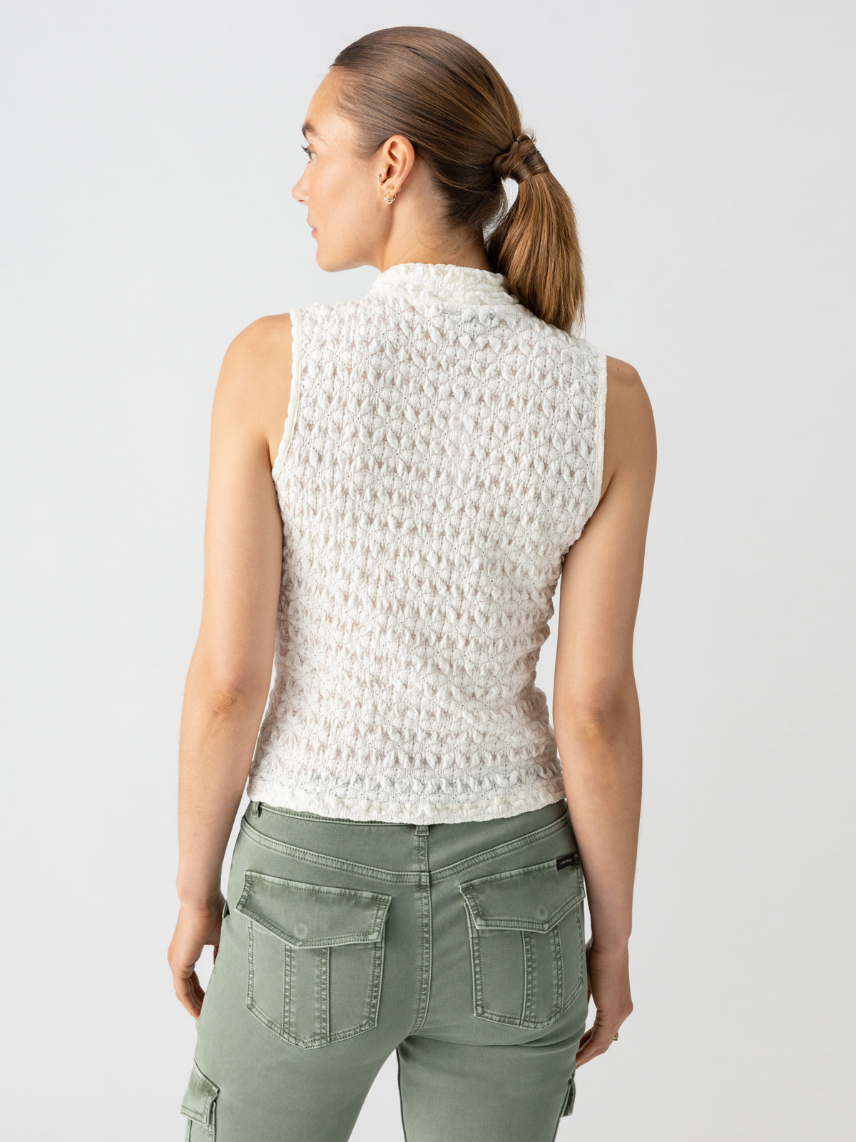 A person with light brown hair in a ponytail is seen from behind, wearing the "Pucker Up Mock Milk" sleeveless white textured top by Sanctuary Clothing, paired with light green cargo pants. They stand against a plain light gray background.