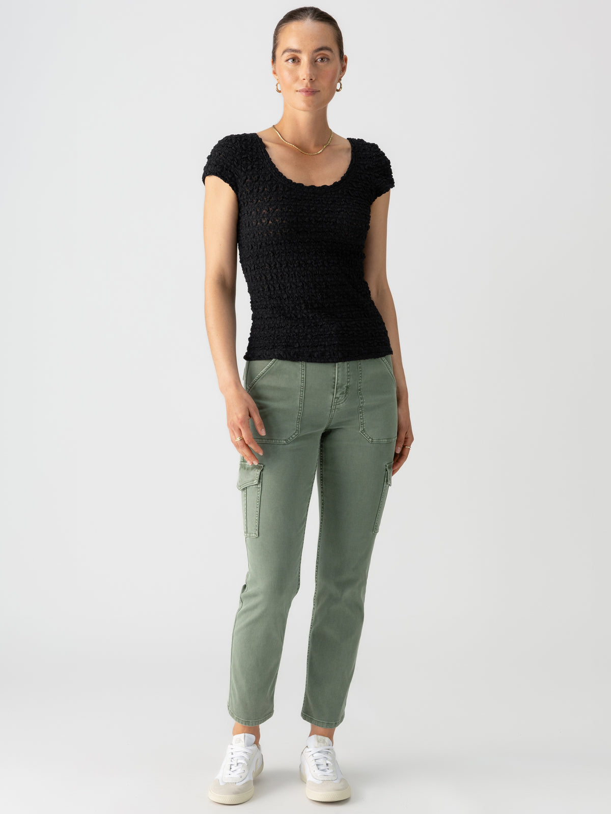 A woman stands against a plain background, wearing the Pucker Up Scoop Cap Tee in black from Sanctuary Clothing paired with sage green cargo pants. She completes her look with white sneakers and has her hair tied back. She looks directly at the camera with a neutral expression.