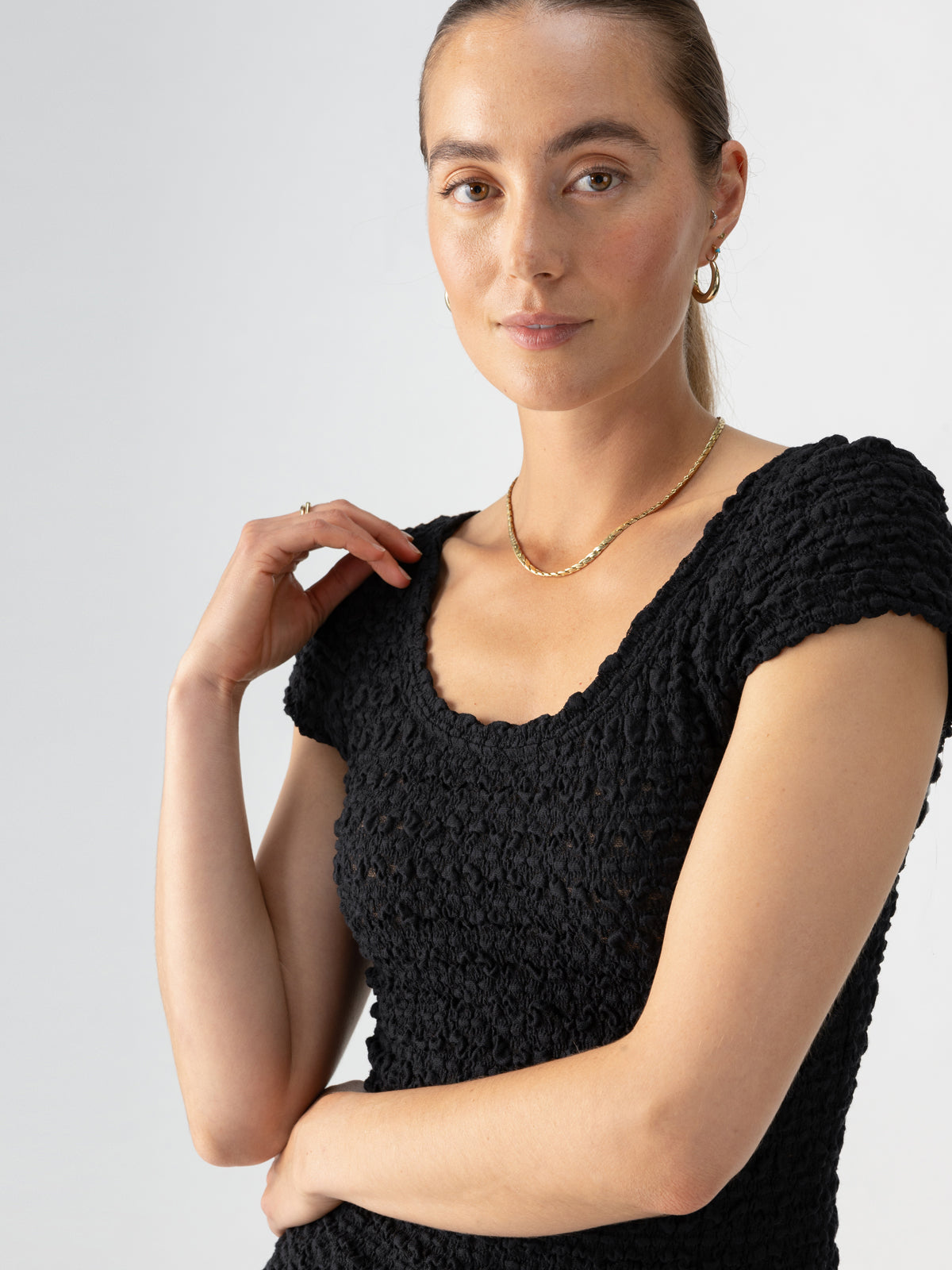 A woman with light skin and brown hair tied back is wearing the "Pucker Up Scoop Cap Tee" in black by Sanctuary Clothing, a gold necklace, and gold hoop earrings. She is standing against a plain background with a relaxed, confident expression, resting one hand near her neck.