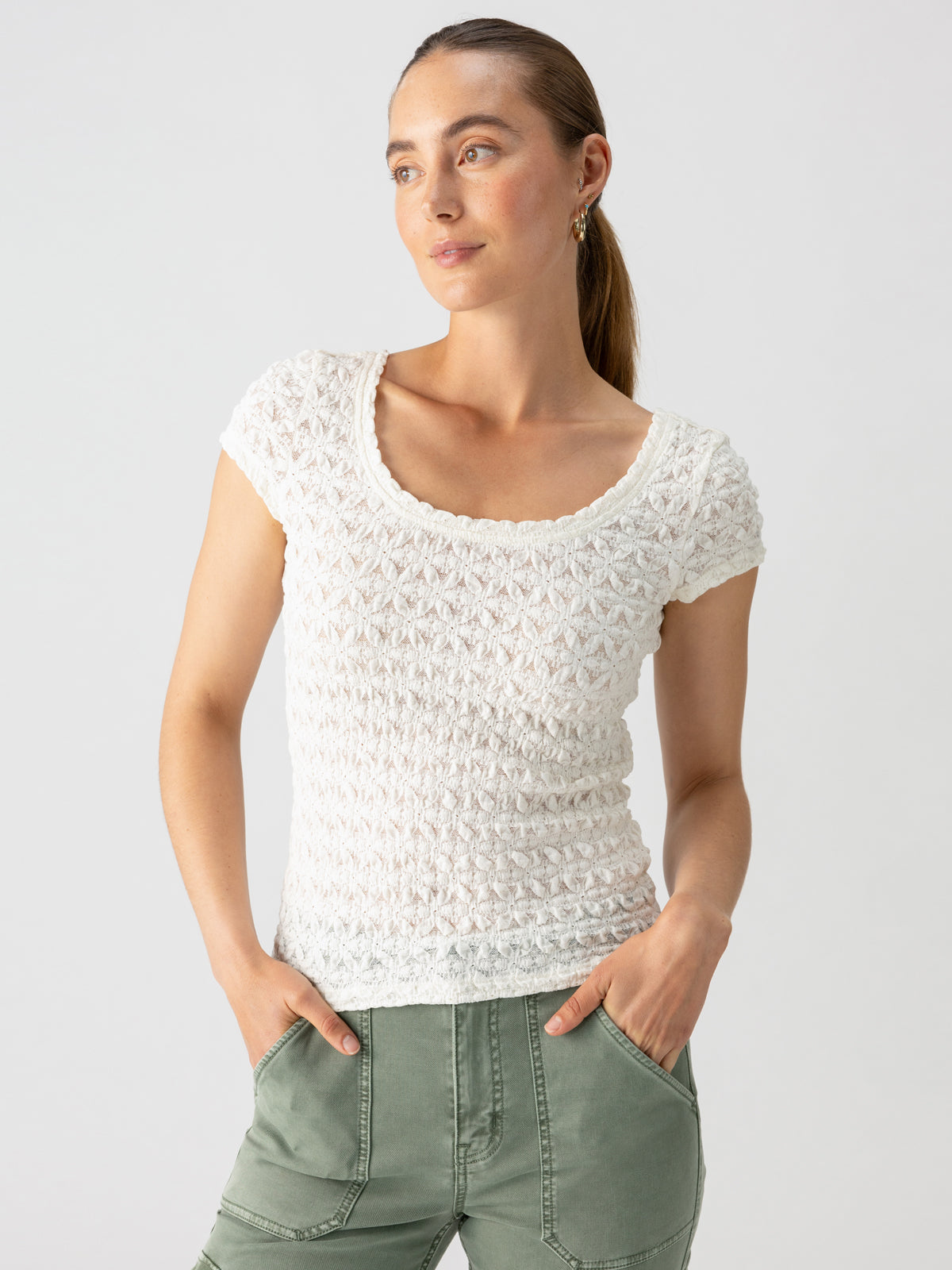 A person with long hair tied back is wearing the Pucker Up Scoop Cap Tee in Milk from Sanctuary Clothing and olive green pants. They are posing with hands in their pockets, looking slightly to the side against a plain light-colored background.